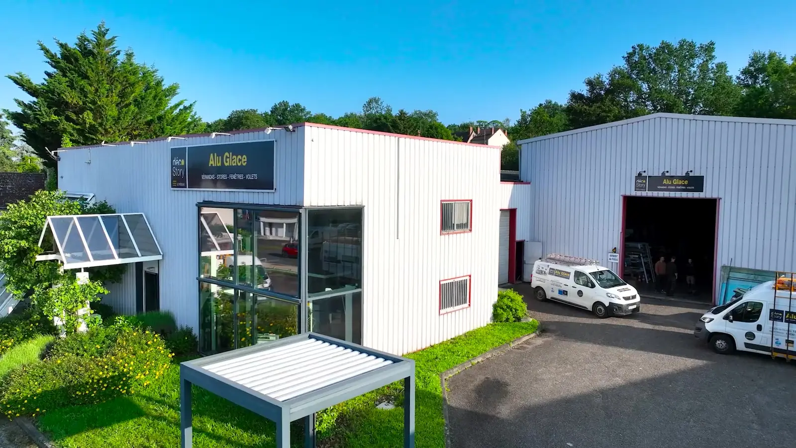 Vue aérienne de l’agence Alu Glace de Vierzon : bâtiment blanc à l’enseigne Alu Glace, entrée vitrée, pergola à lames, camionnettes devant l’atelier