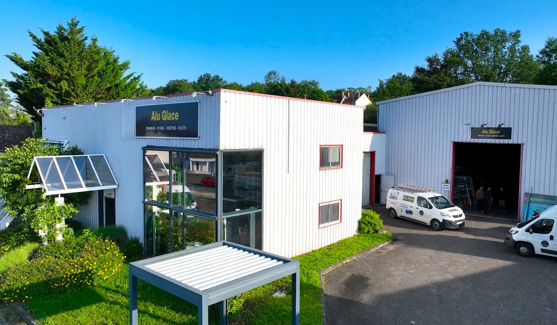 Vue aérienne de l’agence Alu Glace de Vierzon : bâtiment blanc à l’enseigne Alu Glace, entrée vitrée, pergola à lames, camionnettes devant l’atelier
