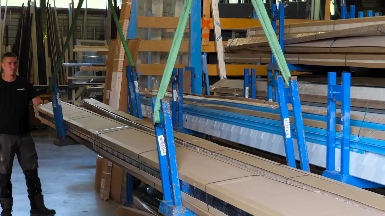 Bottes de profilés soulevées par des sangles vertes devant des rayonnages bleus