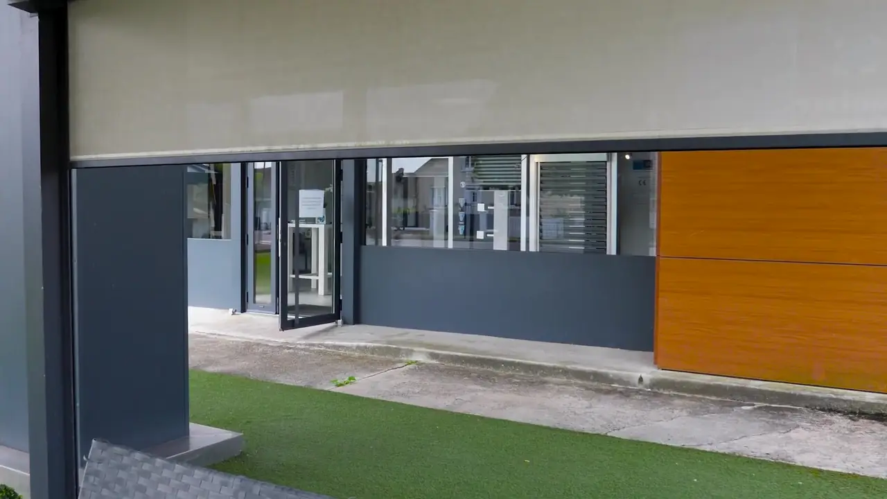 Store vertical gris à demi déroulé entre les poteaux d’une pergola, bâtiment de l’agence derrière