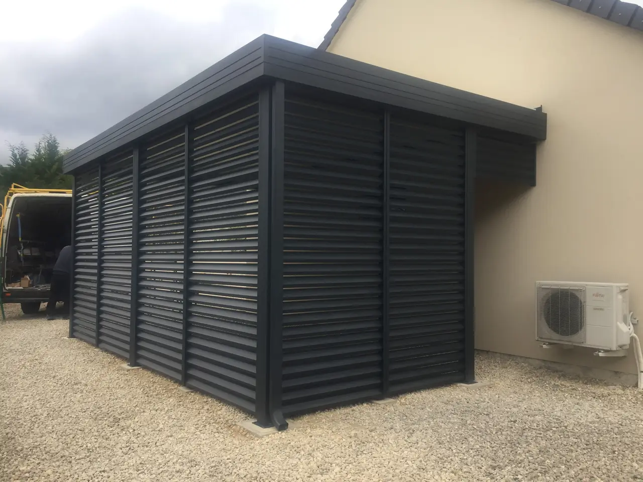 Abri gris anthracite à toit plat et parois de lames horizontales, adossé à une maison