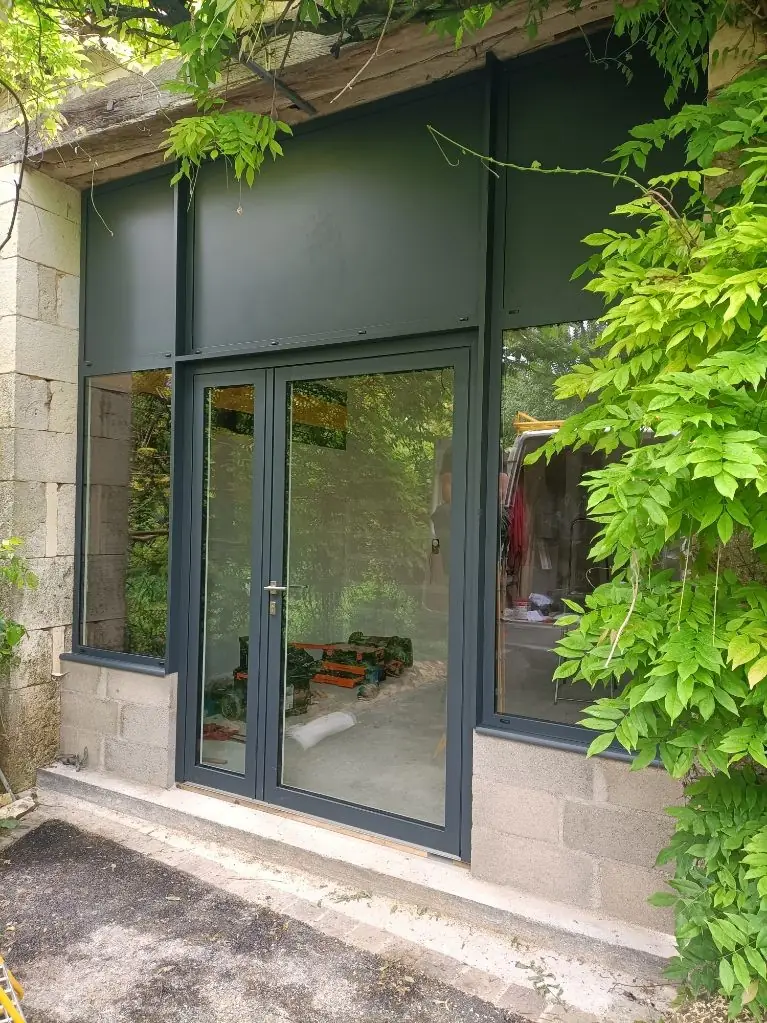 Porte vitrée à deux vantaux et châssis fixes gris anthracite dans un mur de pierre couvert de glycine