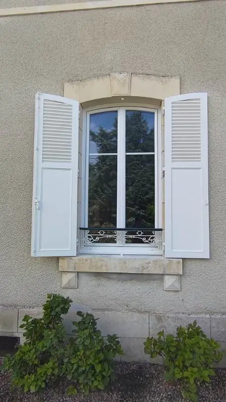 Fenêtre cintrée blanche entre deux volets persiennés blancs, encadrement de pierre