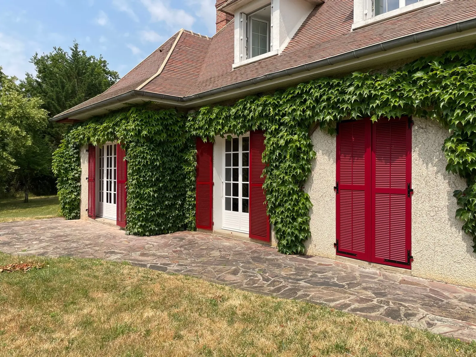 Maison couverte de vigne vierge aux volets persiennés rouges et porte-fenêtres blanches