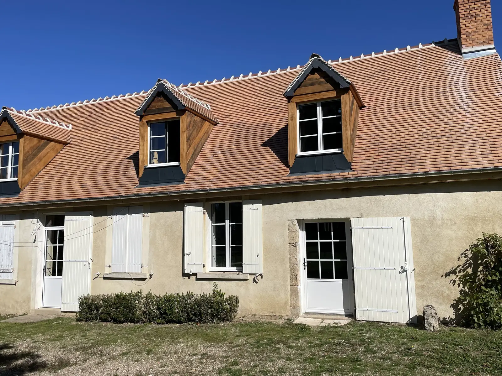 Maison ancienne aux fenêtres, portes et volets battants blancs, deux lucarnes sur le toit de tuiles