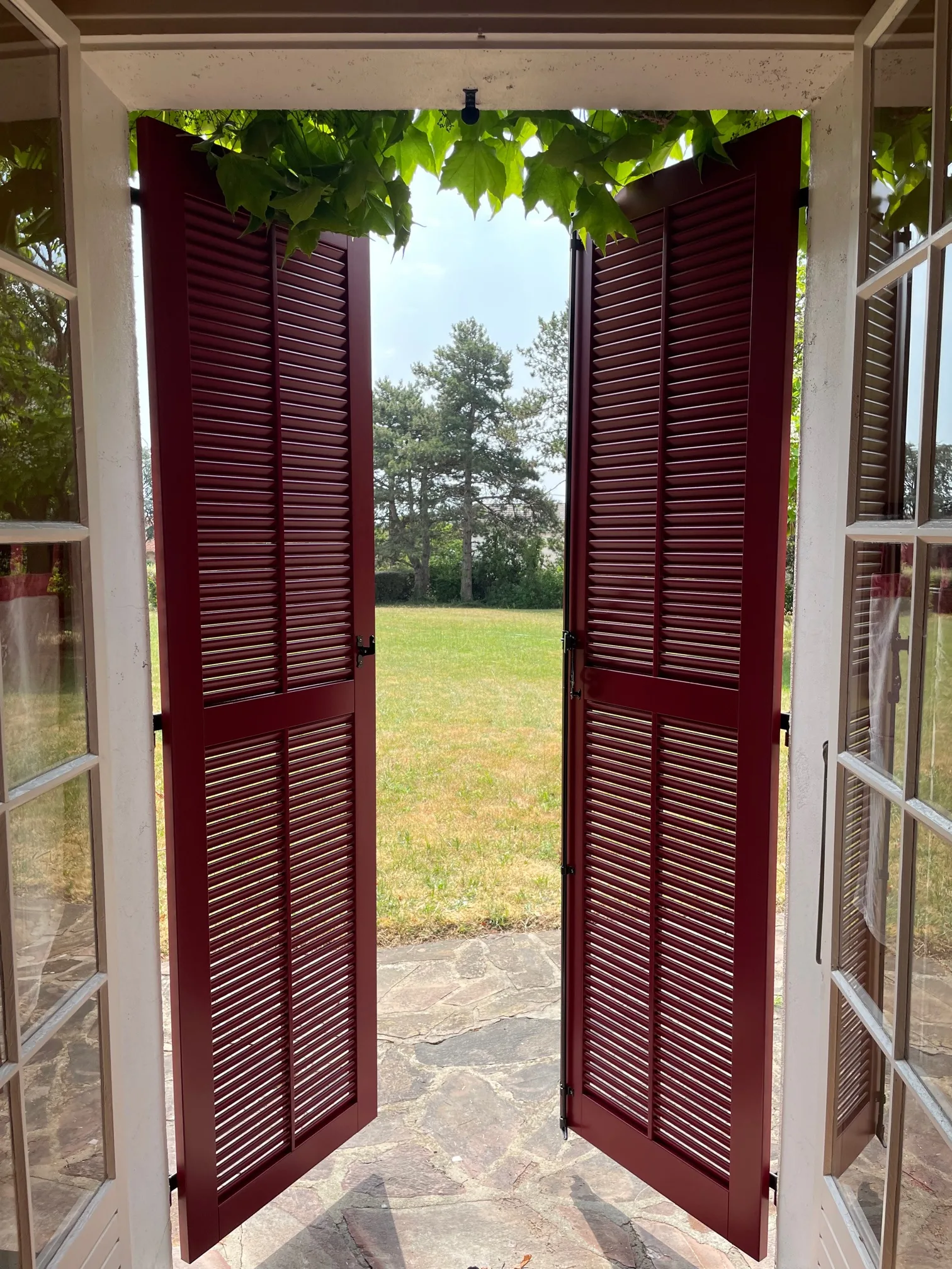Volets persiennés rouges ouverts vus de l’intérieur, terrasse et jardin au fond