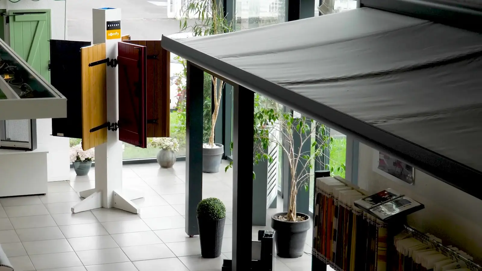 Showroom d’Alu Glace : store banne gris déployé, présentoir de volets battants, plantes et nuanciers de toiles